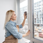 Young Smiling Woman Washing Window with Sponge. Happy Beautiful Girl Cleaning Window by spraying Cleaning Products and wiping with Sponge. Woman Cleaning House.